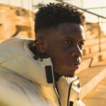 Black teen in a cream coat sitting outside on steps